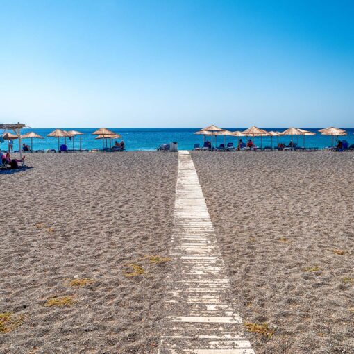 Koutsounari Long Beach neďaleko Ierapetry na juhovýchodnej Kréte ponúka dlhé pásy tmavého piesku, tyrkysové more a pokojnú atmosféru. Pláž lemujú slamené slnečníky a drevený chodník vedúci priamo k vode, ideálne miesto na oddych mimo davov.